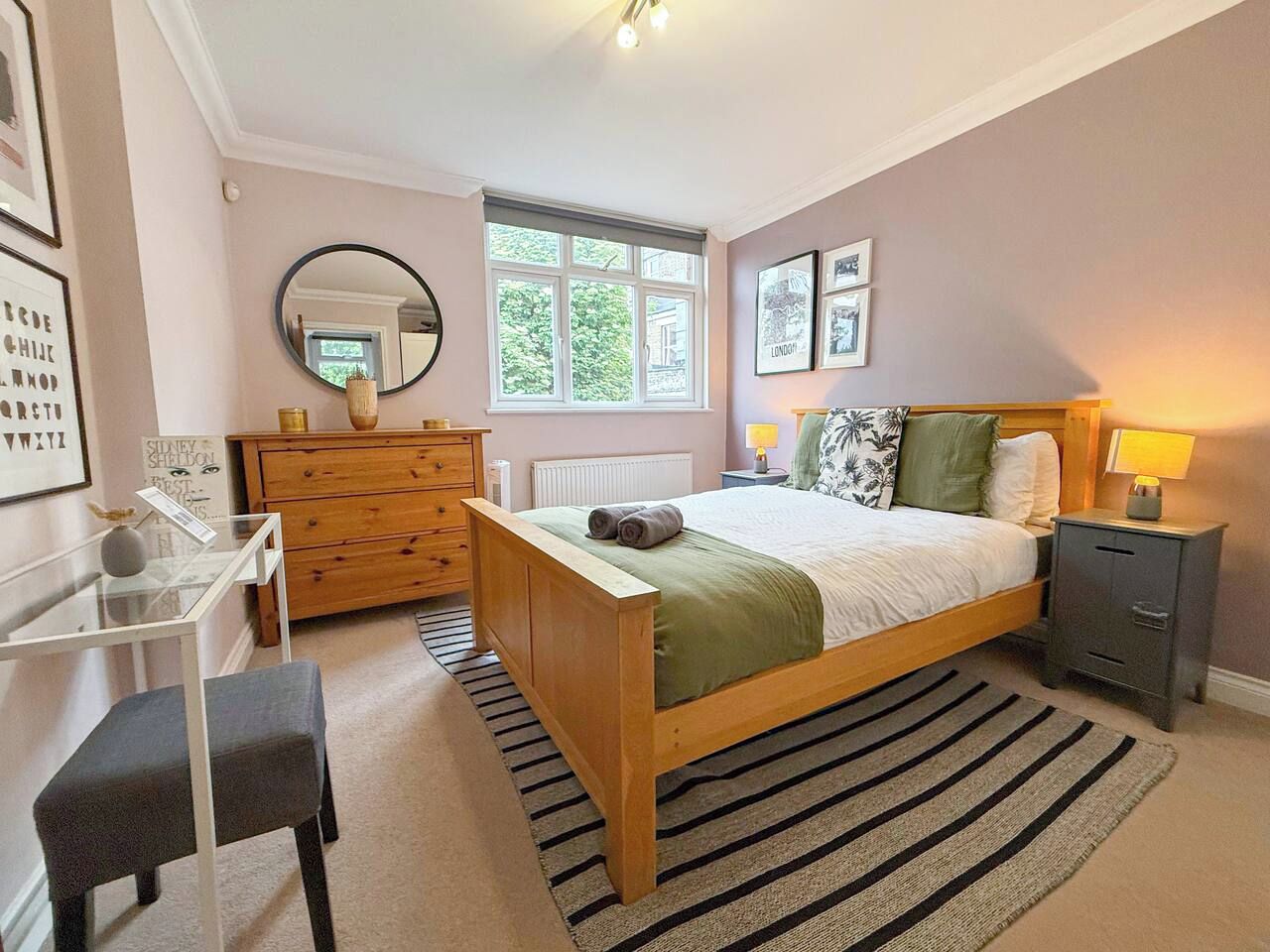 A bedroom with a bed, bedside table, dresser, mirror, and a window.