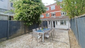 Un'area patio con tavolo e sedie, circondata da un'alta recinzione, e un edificio con tetto in tegole sullo sfondo.