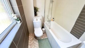 Een kleine badkamer met toilet, bad en een klein raam met een plantje.