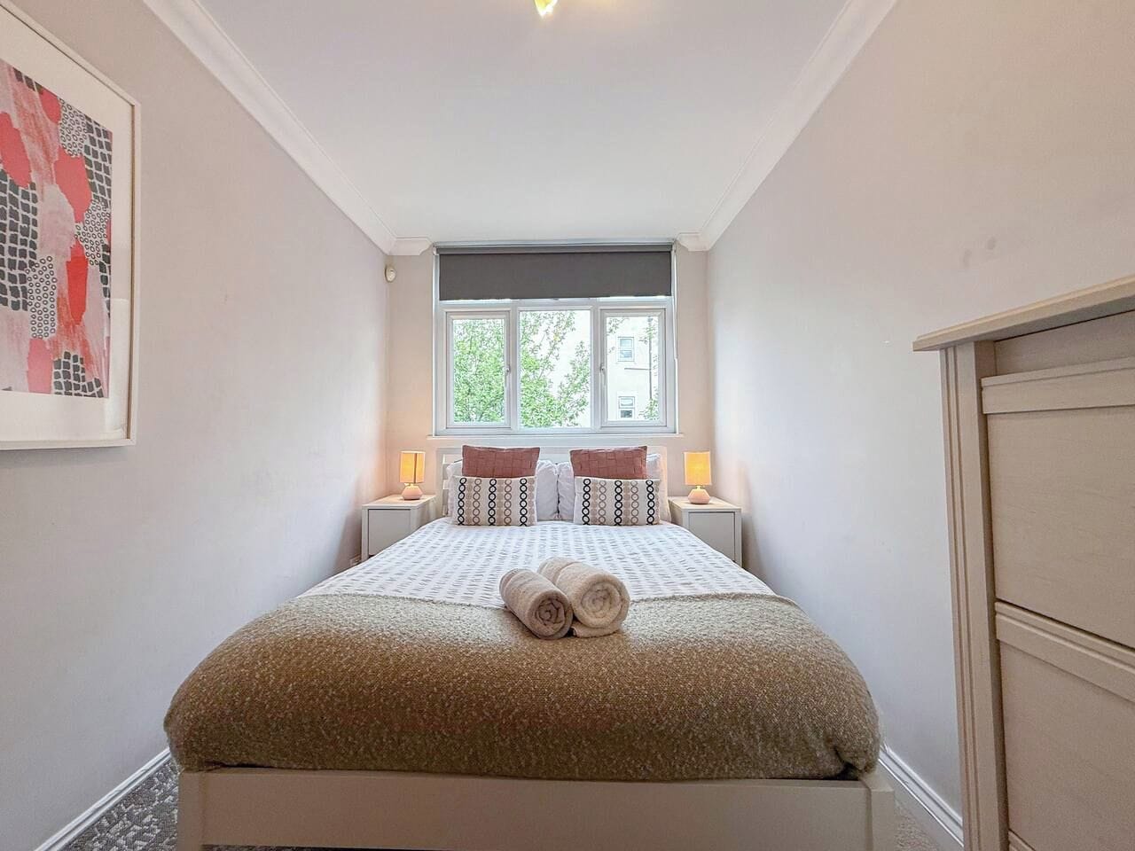 A bedroom with a bed, two bedside lamps, a window, and a painting on the wall.