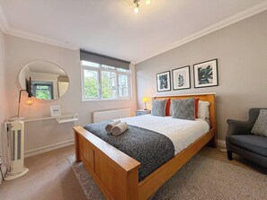 Een slaapkamer met een bed, nachtkastje, lamp, radiator, raam en een stoel.
