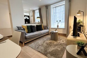A modern living room with a grey sofa, a round coffee table, a small side table, a TV stand, and a large window with curtains.