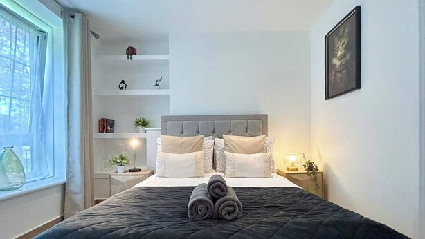 Cama bem arrumada com colcha escura e toalhas dobradas, mesa de cabeceira com abajur, prateleira com objetos decorativos e janela com cortinas.