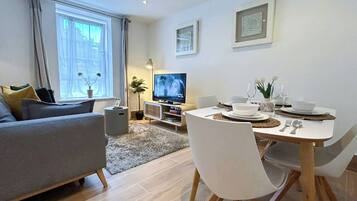 A modern living room with a sofa, a dining table set for two, a television, and a potted plant.