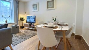Uma sala de jantar moderna com uma mesa redonda posta para dois, uma televisão ao fundo e um sofá em primeiro plano.