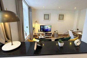 A modern living room with a flat-screen TV, a dining area with a table set for two, and a large window with curtains.