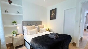 Quarto com cama, mesas de cabeceira, um criado-mudo, um quadro na parede e uma porta.