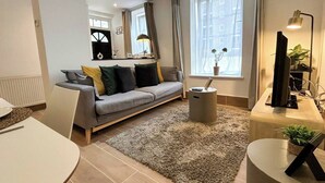 Uma sala de estar moderna com um sofá cinzento, uma mesa de centro redonda, um rack para TV e uma janela grande com cortinas.