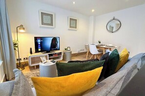 A modern living room with a flat-screen TV, a dining area with a table and chairs, and a sofa with cushions.