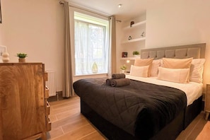 A bedroom with a large bed, a wooden dresser, a window with curtains, and a bedside table with a lamp.