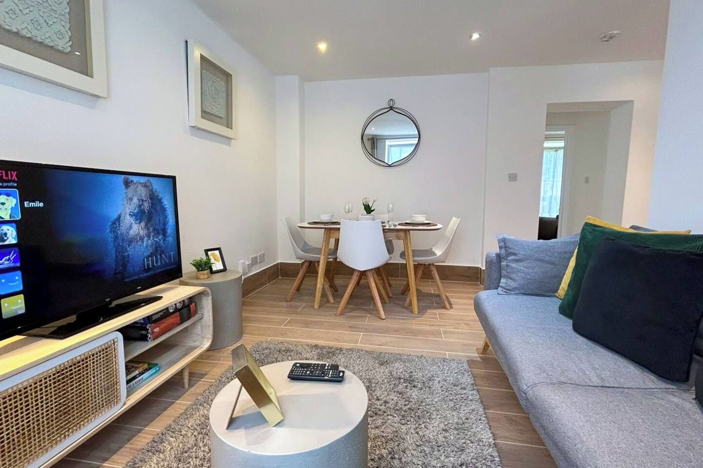 A modern living room with a flat-screen TV, a dining area with a round table and chairs, a grey sofa, and a small round coffee table.