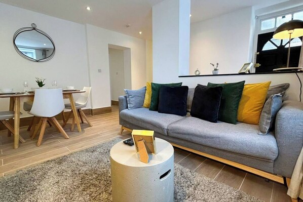 A modern living room with a grey sofa, a dining table with chairs, a round coffee table, and a mirror on the wall.