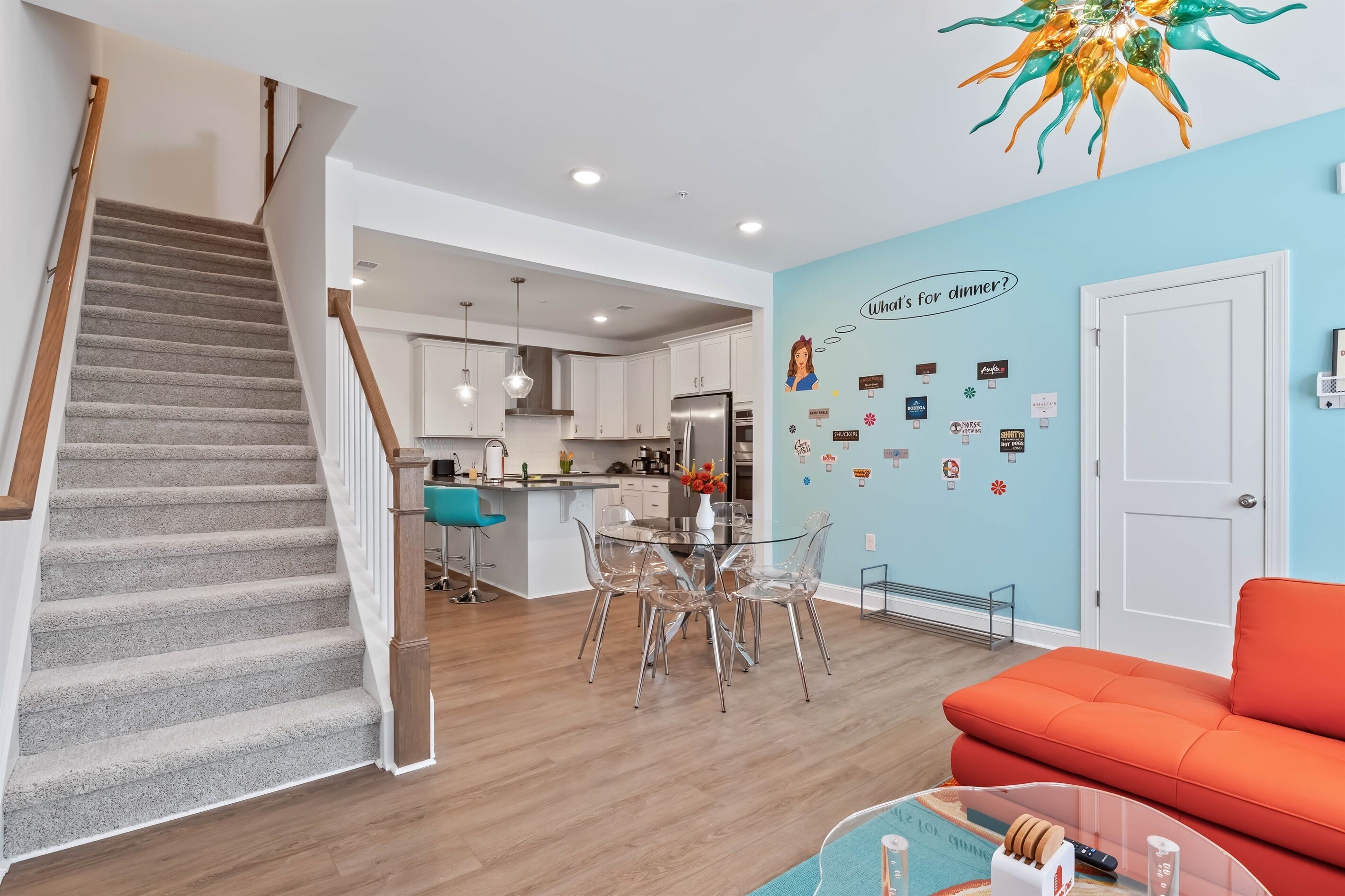 A modern kitchen with a dining area, a staircase, and a living room with an orange sofa.