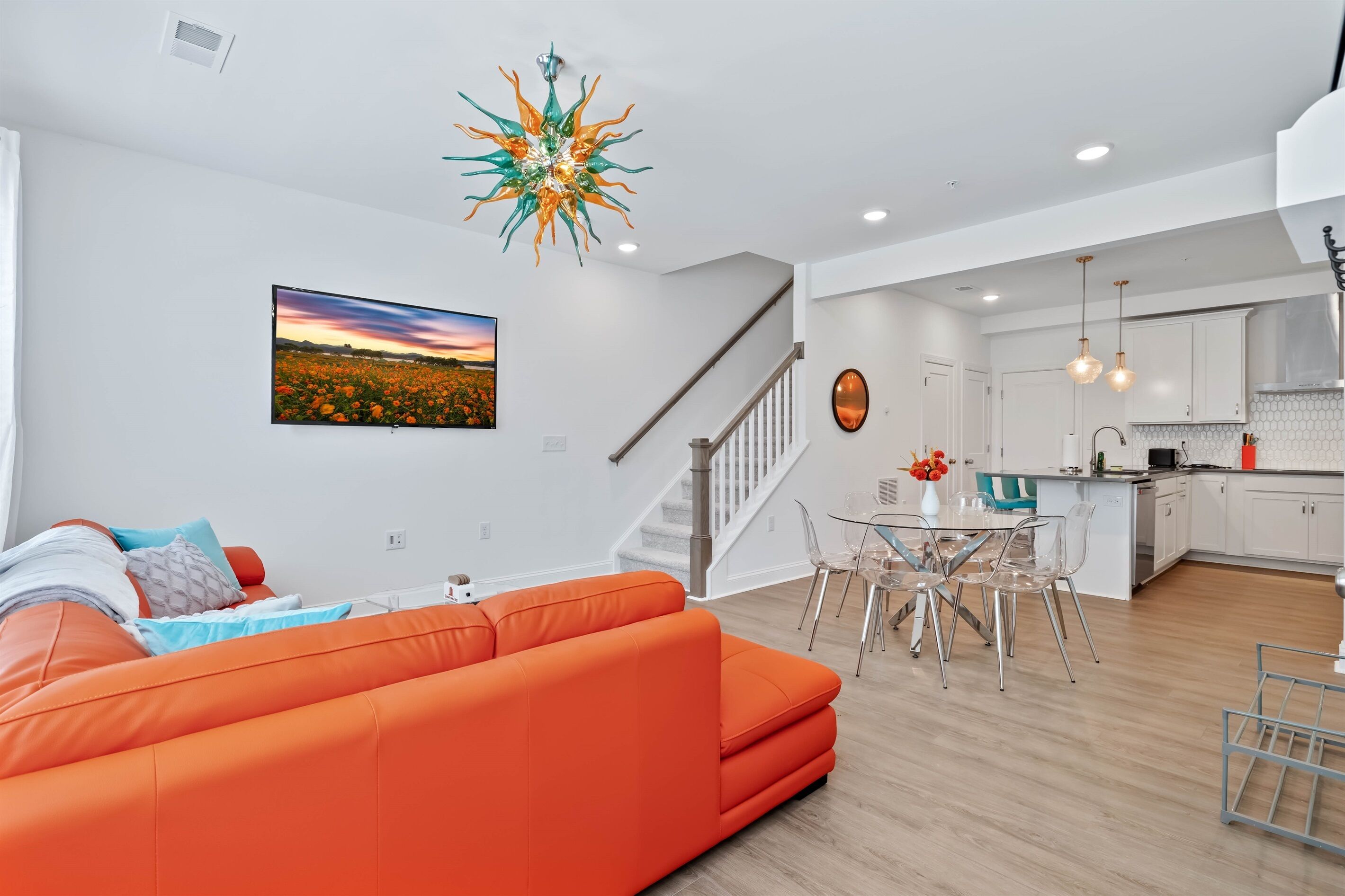 A modern living room with a large orange sofa, a dining area with a glass table, and a kitchen with white cabinets and stainless steel appliances.