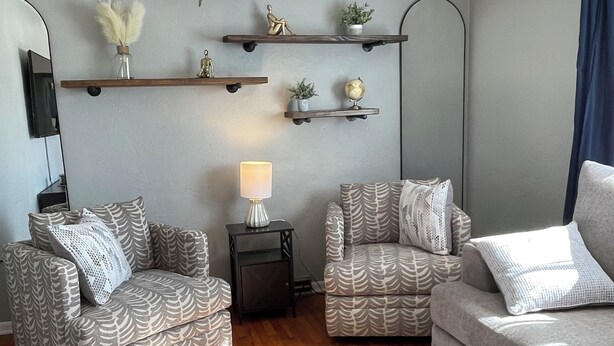 A living room with two patterned armchairs, a sofa, a coffee table, and a TV on the wall. Shelves with decorative items are mounted on the wall. - Charming 3 Bedroom House away from it all but Close to everything (Odessa)