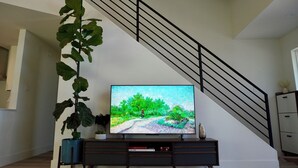 Una sala de estar con un televisor de pantalla plana sobre un mueble bajo de madera oscura, una escalera con barandillas negras y una planta verde grande.