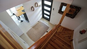 A high-ceilinged room with a wooden staircase, a kitchen area with a dining table, and a door with a mirror and wall decorations. - La Villa de Font-Romeu (Font-Romeu-Odeillo-Via)
