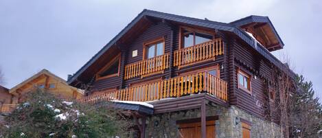 A log cabin with a stone foundation, wooden balcony, and a stone wall in front.