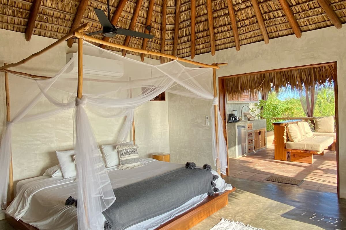 A room with a wooden four-poster bed, a thatched roof, and a view of the outdoors.
