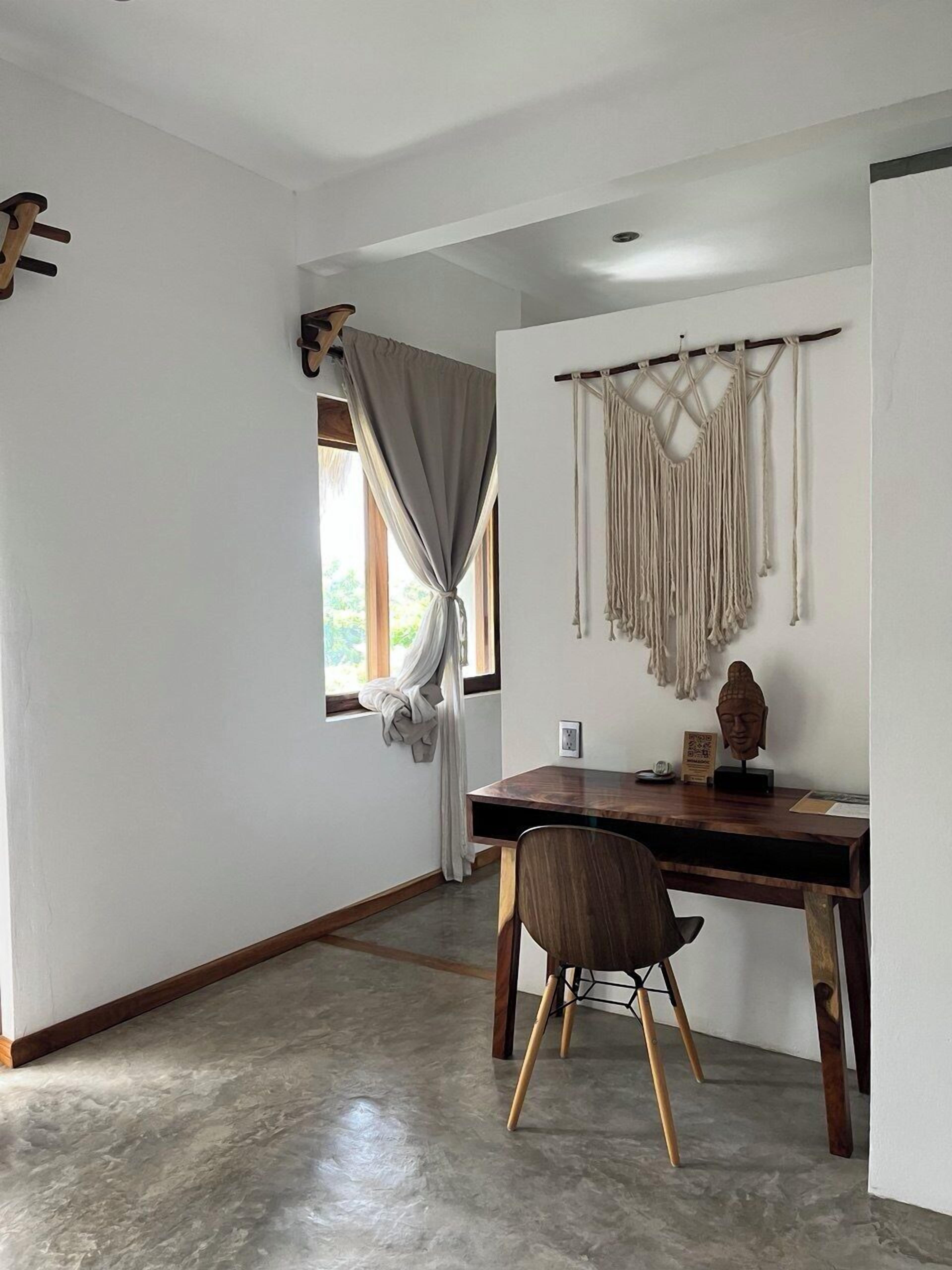 Una habitación con escritorio de madera, una silla, un tapiz de macramé y una ventana con cortinas.