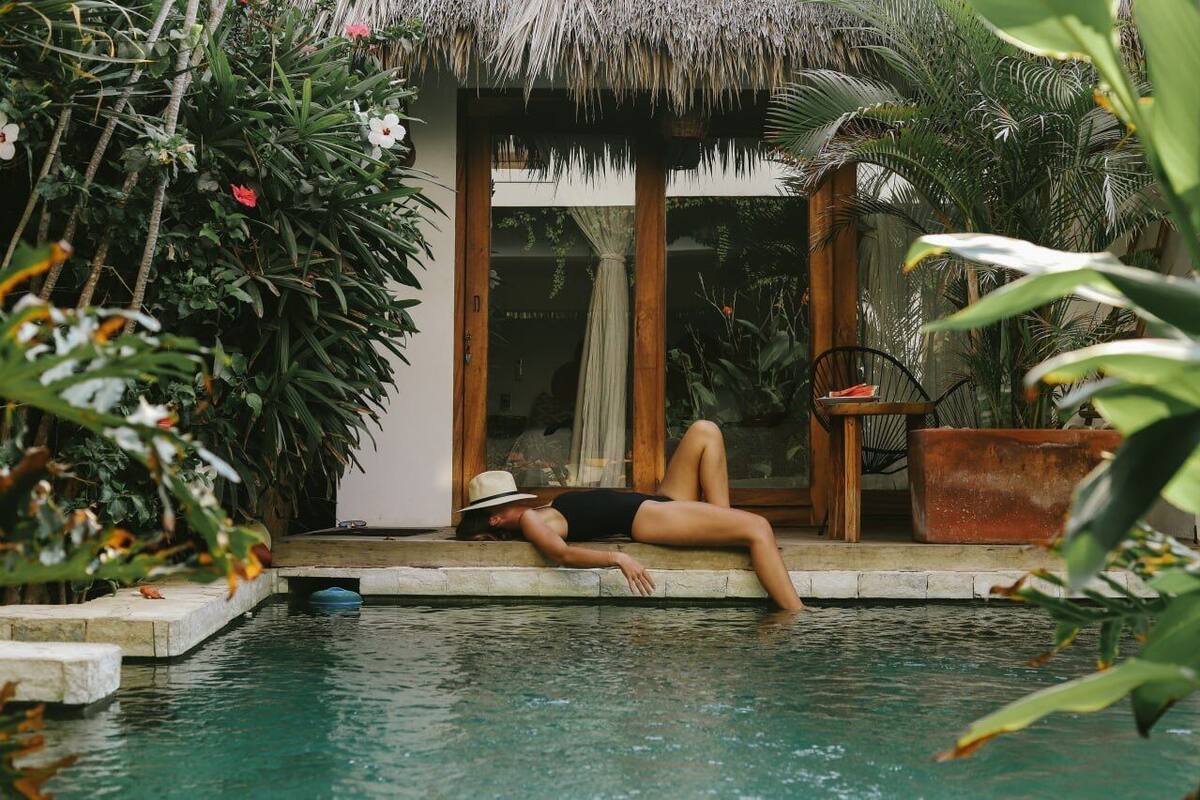 A person relaxing by a poolside with a thatched roof structure and lush greenery in the background.