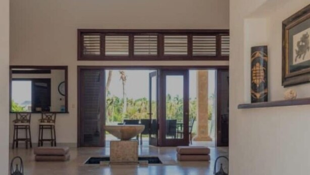 A spacious interior with a high ceiling, a central seating area, and a view through large glass doors to an outdoor space.