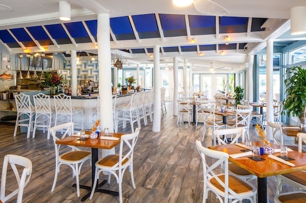 A spacious restaurant with high ceilings, white chairs, and wooden tables.