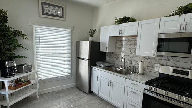 A modern kitchen with white cabinets, a stainless steel refrigerator, a microwave, a stove, and a sink. There is a window with blinds and a plant on the counter.