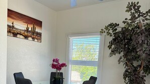 A dining area with a glass table, four chairs, a ceiling fan, a painting, a window with blinds, and a potted plant.