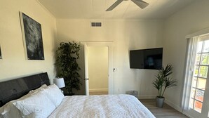 A bedroom with a large bed, a ceiling fan, a TV, and a door leading to another room.