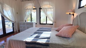 A bedroom with a bed, a bedside lamp, a window with curtains, and a view of the outside. - 10 Minute Walk to Pickleball and the Sea of Cortez (Los Barriles)