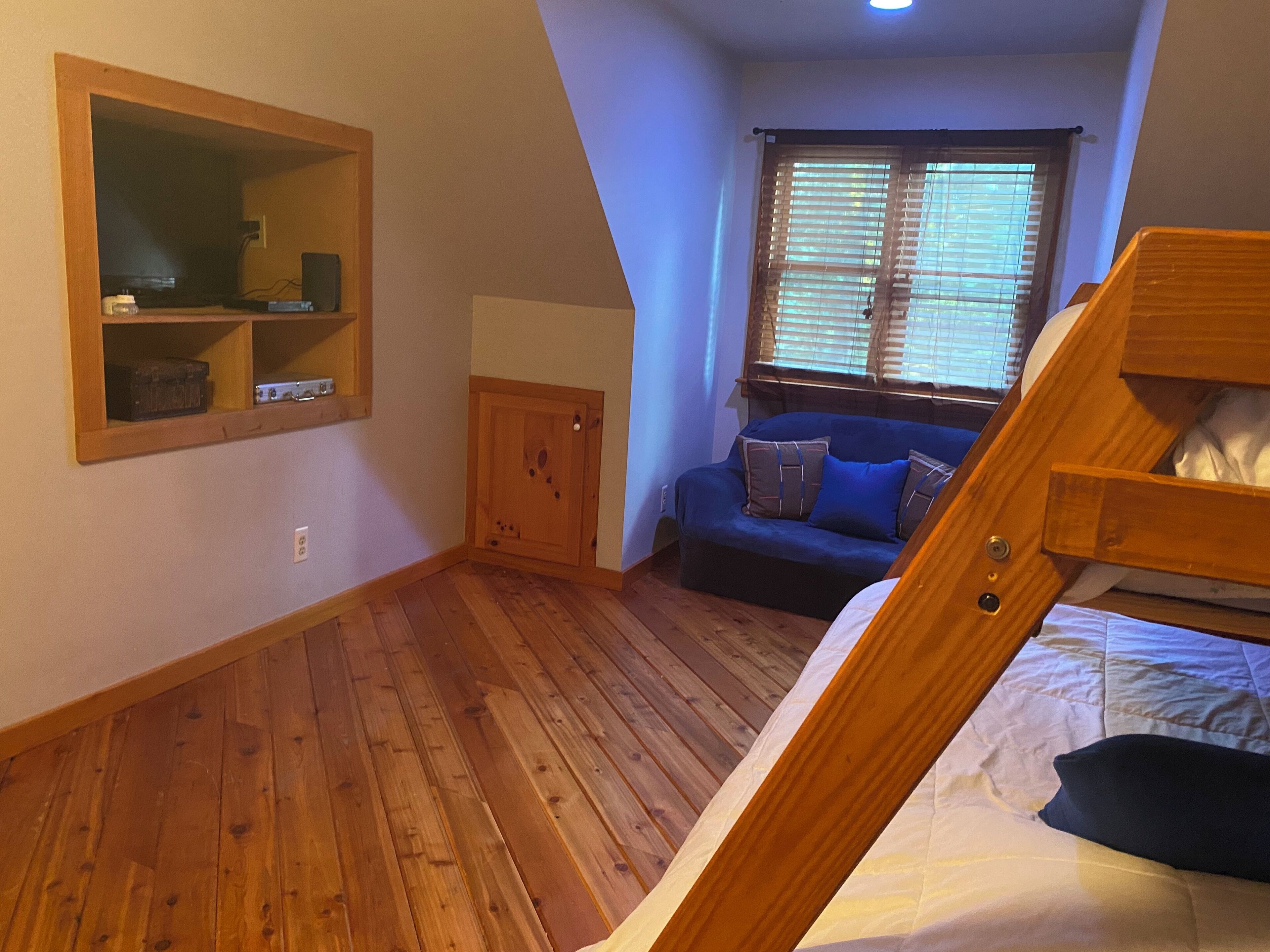 A bedroom with a wooden floor, a bunk bed, a sofa with cushions, a wooden cabinet, and a window with blinds.