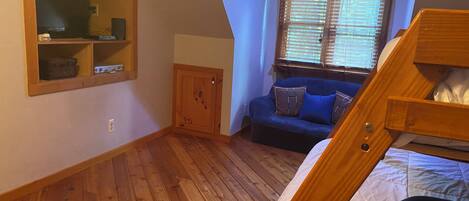 A bedroom with a wooden floor, a bunk bed, a sofa with cushions, a wooden cabinet, and a window with blinds.