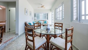 A dining area with a round glass table surrounded by wicker chairs, a ceiling fan, and large windows with shutters.