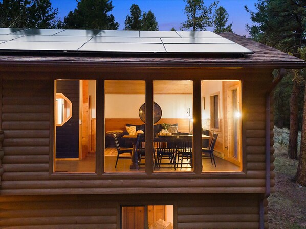 Une cabane en rondins avec un coin repas visible à travers de grandes fenêtres, une façade en bois et une vue sur les arbres en arrière-plan.