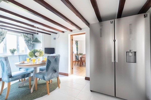 A modern kitchen with a stainless steel refrigerator, wooden beams on the ceiling, and a dining area with blue chairs and a round table. - Beautiful Danford House & Lodge with Hot Tub (Hartpury)