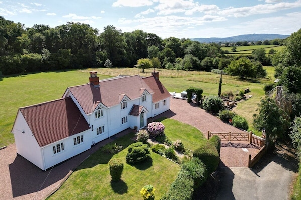 A large white house with a red roof, surrounded by a well-maintained lawn and garden, with a view of rolling hills in the background.