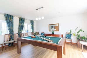 Una mesa de billar en una sala con suelo de madera, sillas y un amplio ventanal con cortinas.