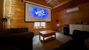 Quarto aconchegante com paredes de madeira, uma tela de projetor exibindo 'Disney Pixar Inside Out 2', uma mesa pequena e dois sofás.