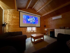 Una habitación acogedora con paredes de madera, una pantalla de proyector mostrando 'Disney Pixar Inside Out 2', una mesita pequeña y dos sofás.