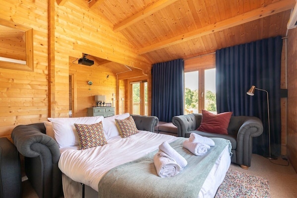 A wooden cabin room with a bed, two armchairs, a desk, and a window with curtains.