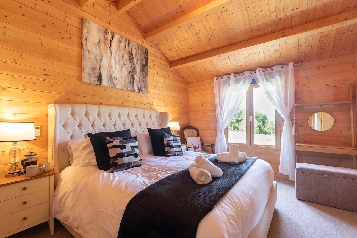 A wooden cabin bedroom with a large bed, a nightstand, a bedside table, a window with curtains, and a wooden wall with a painting.
