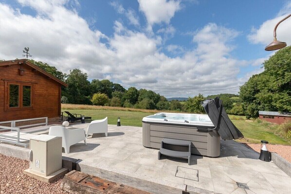 Un jacuzzi con tapa, mobiliario de exterior y una cabaña de madera en una zona ajardinada.