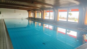 An indoor swimming pool with clear blue water, wooden beams on the ceiling, and large windows with red awnings.