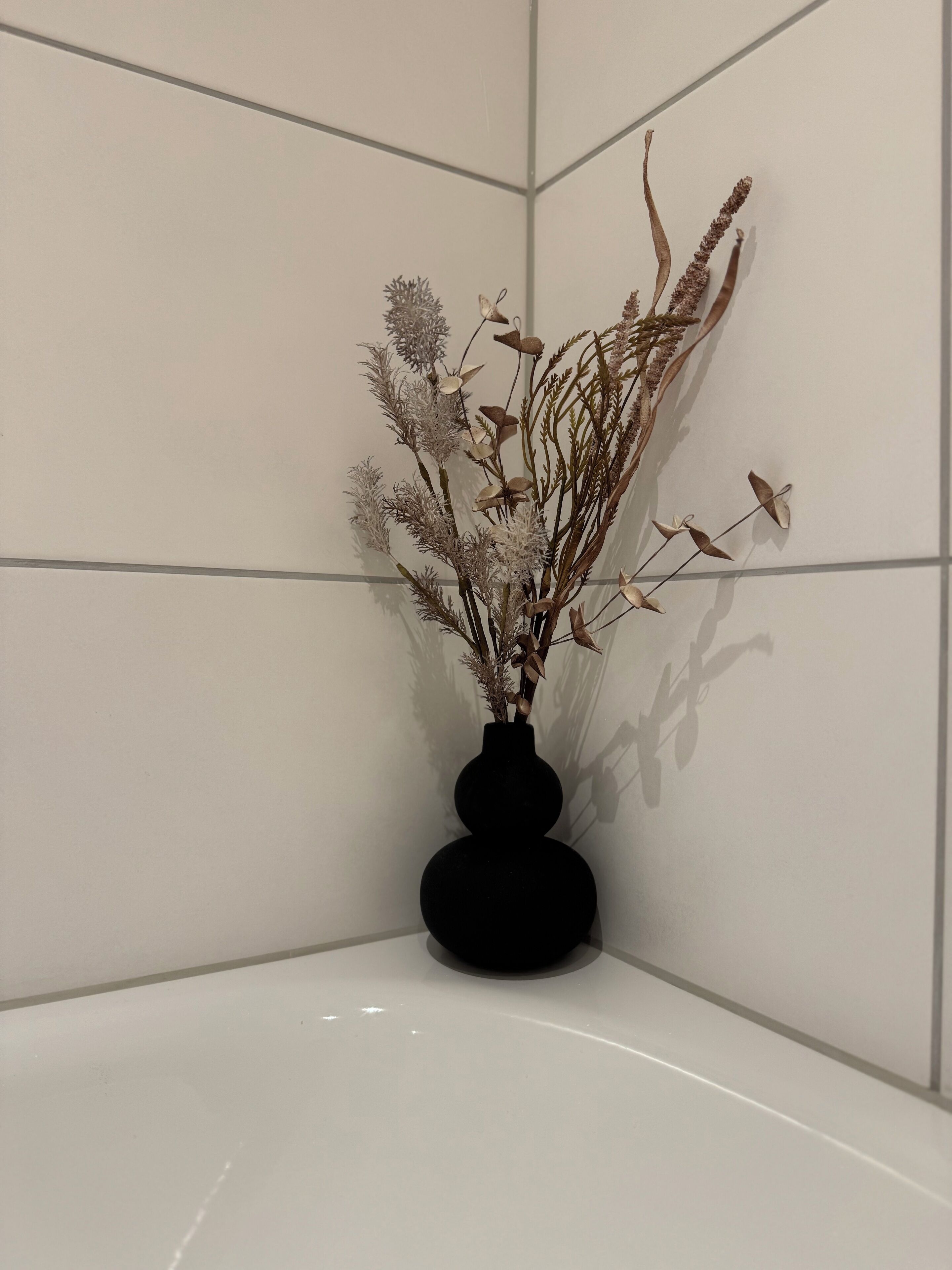 Una cantonada d'una habitació amb parets de rajola blanca i un vas negre amb plantes seques.