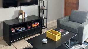 A living room with a flat-screen TV, a grey sofa, a coffee table, a black entertainment unit, and a door. - Charming 3-bedroom house with AC, WiFi in phenomenal Phoenix (Phoenix)