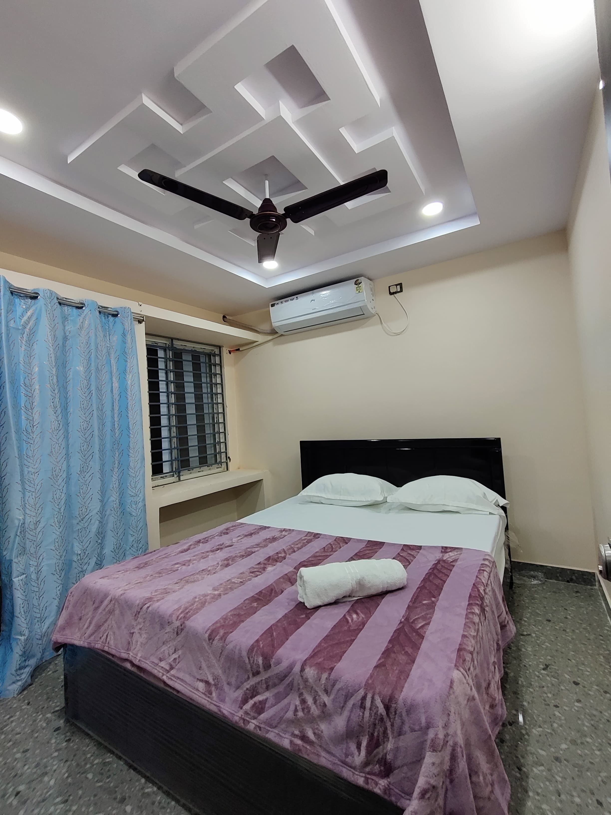 A bedroom with a bed, a ceiling fan, an air conditioning unit, a window with bars, and a curtain.