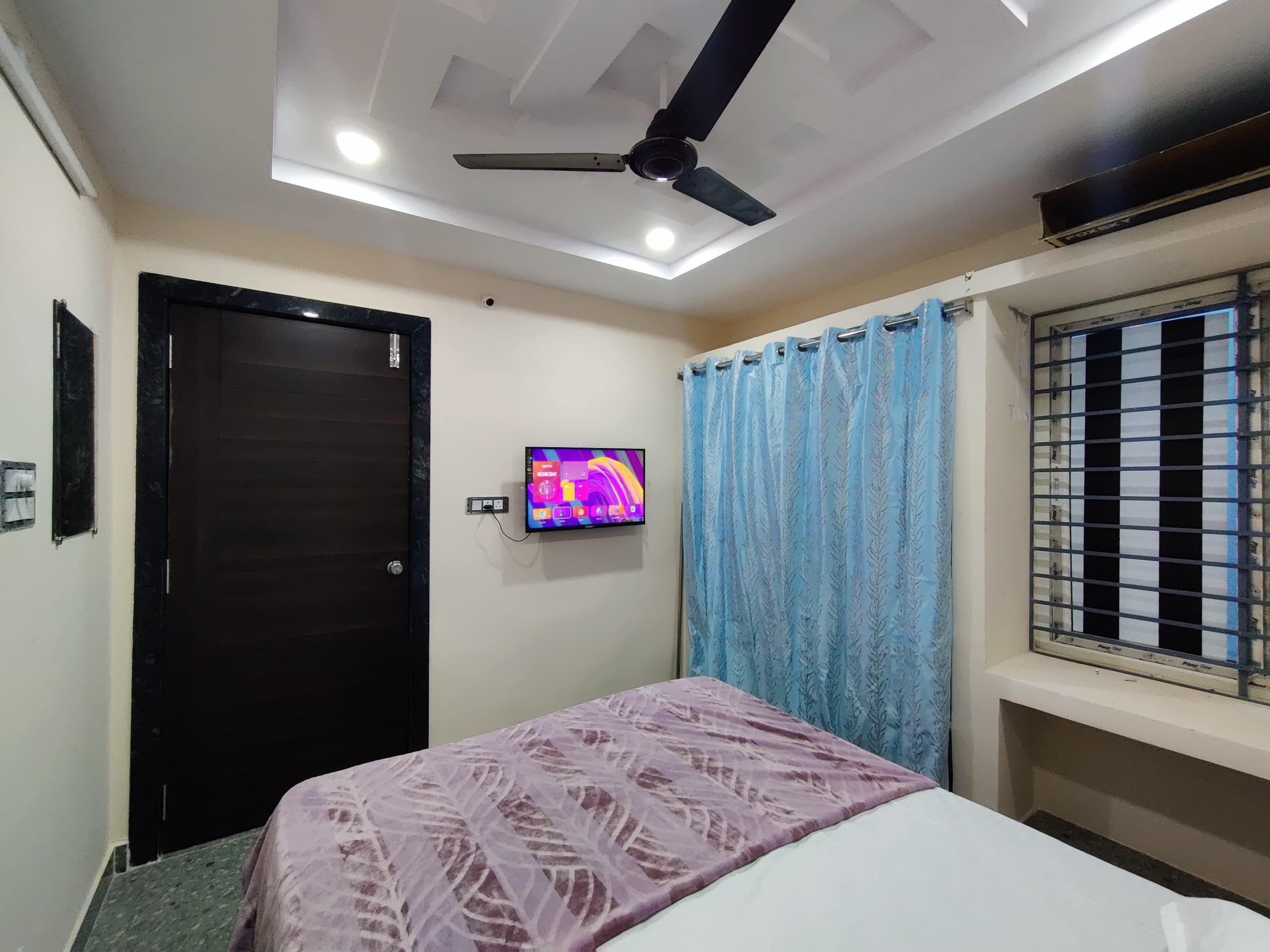 A bedroom with a bed, a ceiling fan, a television, and a window with bars.
