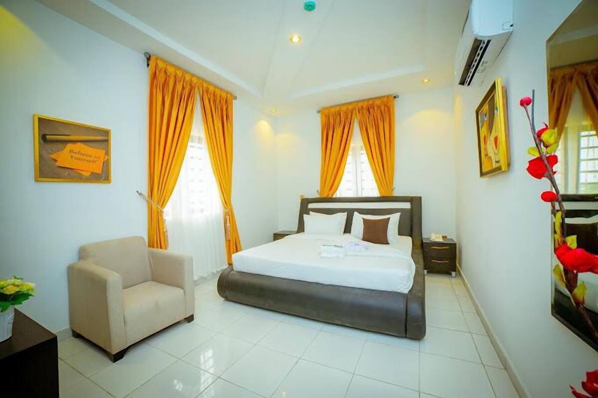 A hotel room with a bed, a chair, a nightstand, and a wall-mounted air conditioner.