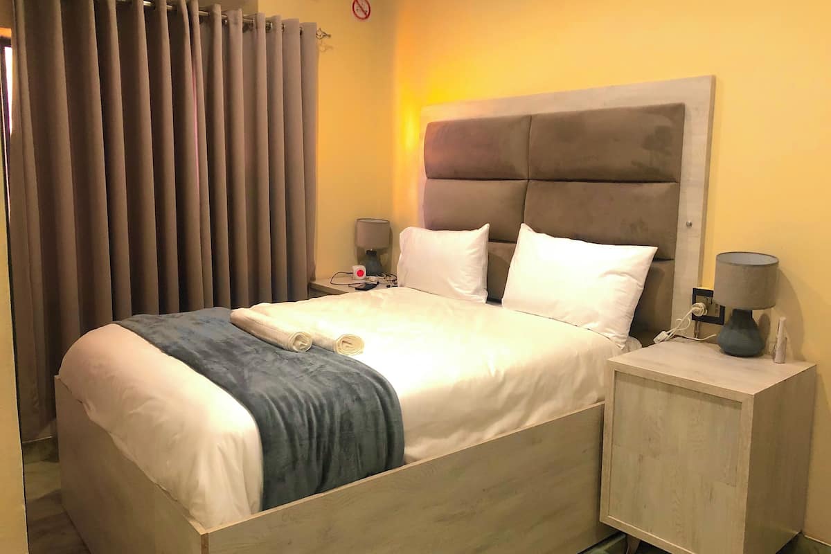 Una cama bien hecha con cabecera, dos mesitas de noche con lámparas, una ventana con cortinas y una lámpara de pared.